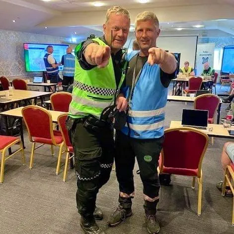 To menn i uniform og refleksvest peker mot kameraet i et møterom under en samling