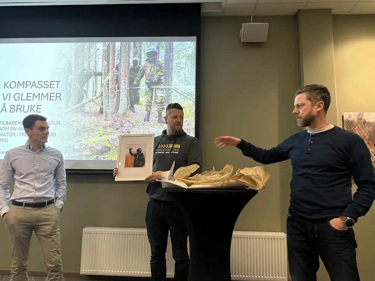 Foredrag og overrekkelse av et innrammet bilde under et arrangement med tre personer foran lerretet
